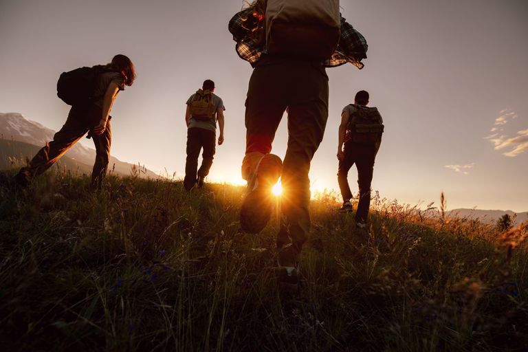 Silhouettes of four young hikers with backpacks are walking in mountains at sunset time